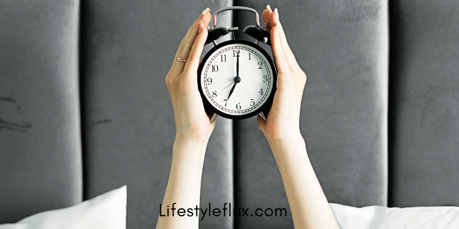 A hand with a ring on the finger is reaching to tap a smartphone screen displaying an alarm set for 4:00 AM. The background shows a bed with a headboard, and the phone is placed on a bedside table. The website "lifestylelux.com" is visible in the bottom left corner.
