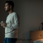 A man holding a mug, standing by a window in a dimly lit bedroom, gazing outside in the early morning light, suggesting a calm and mindful start to the day.