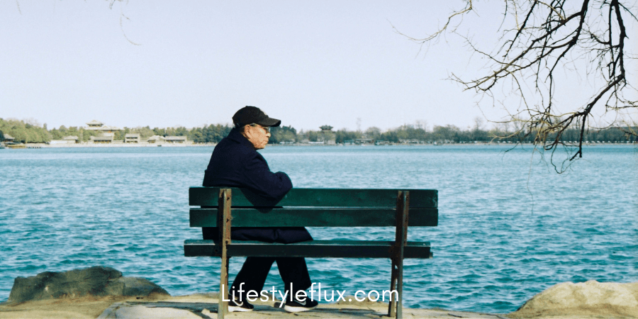 A person wearing a dark jacket and cap is sitting alone on a park bench facing a calm body of water.