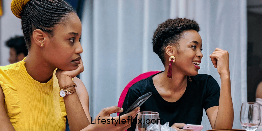 Two young women are sitting at a table, one looking thoughtful while the other is speaking animatedly and holding a phone.