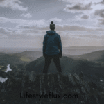 A person in a hoodie and beanie stands on a mountain peak, looking out over a vast, cloudy landscape with valleys and rivers.