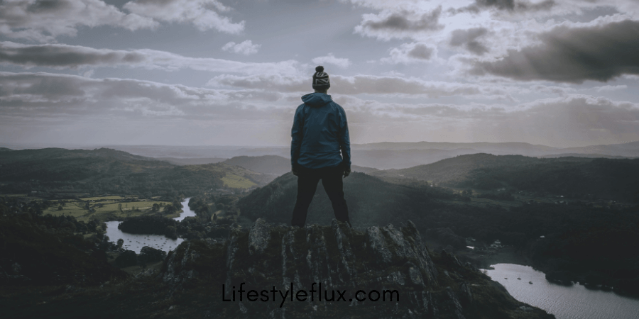 A person in a hoodie and beanie stands on a mountain peak, looking out over a vast, cloudy landscape with valleys and rivers.