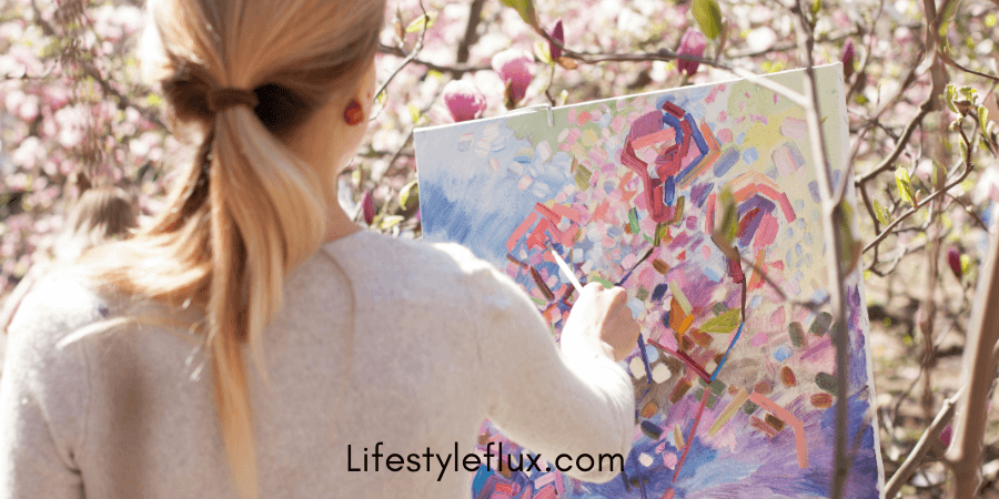 A person with a ponytail, viewed from behind, paints on a canvas outdoors amidst blooming pink flowers.