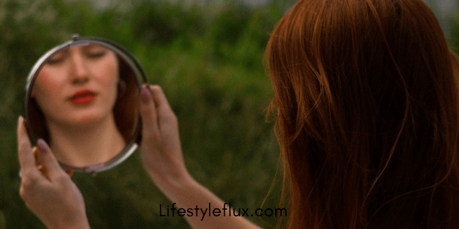 A person with reddish-brown hair holds up a small, round mirror, looking at their reflection.